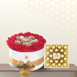 Round white flower box containing red roses surrounding gold roses, tied with a gold ribbon, next to a large box of Ferrero Rocher. Main image