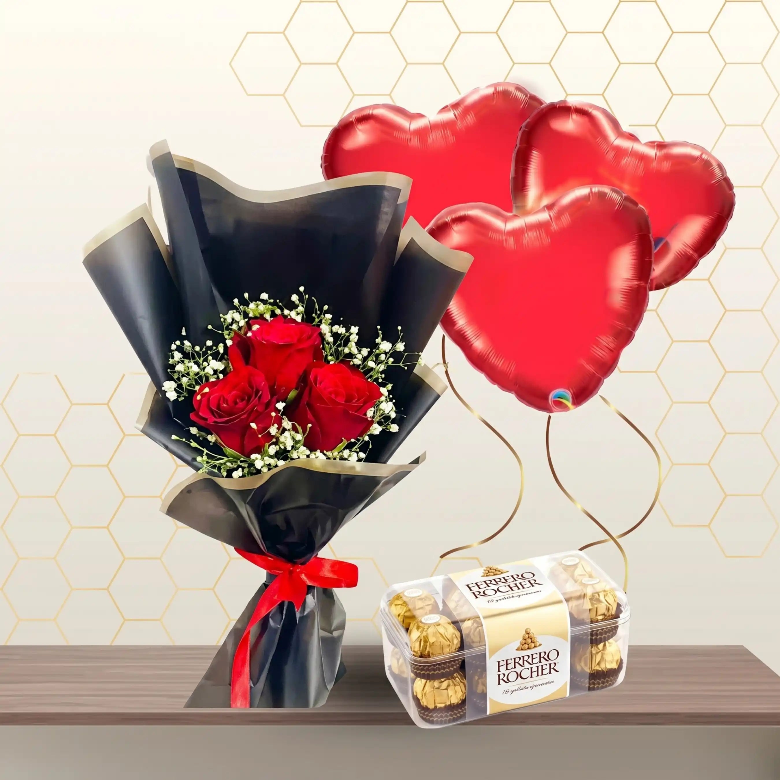 Small bouquet of red roses and baby's breath in black wrapping, next to a box of chocolates and three heart balloons.