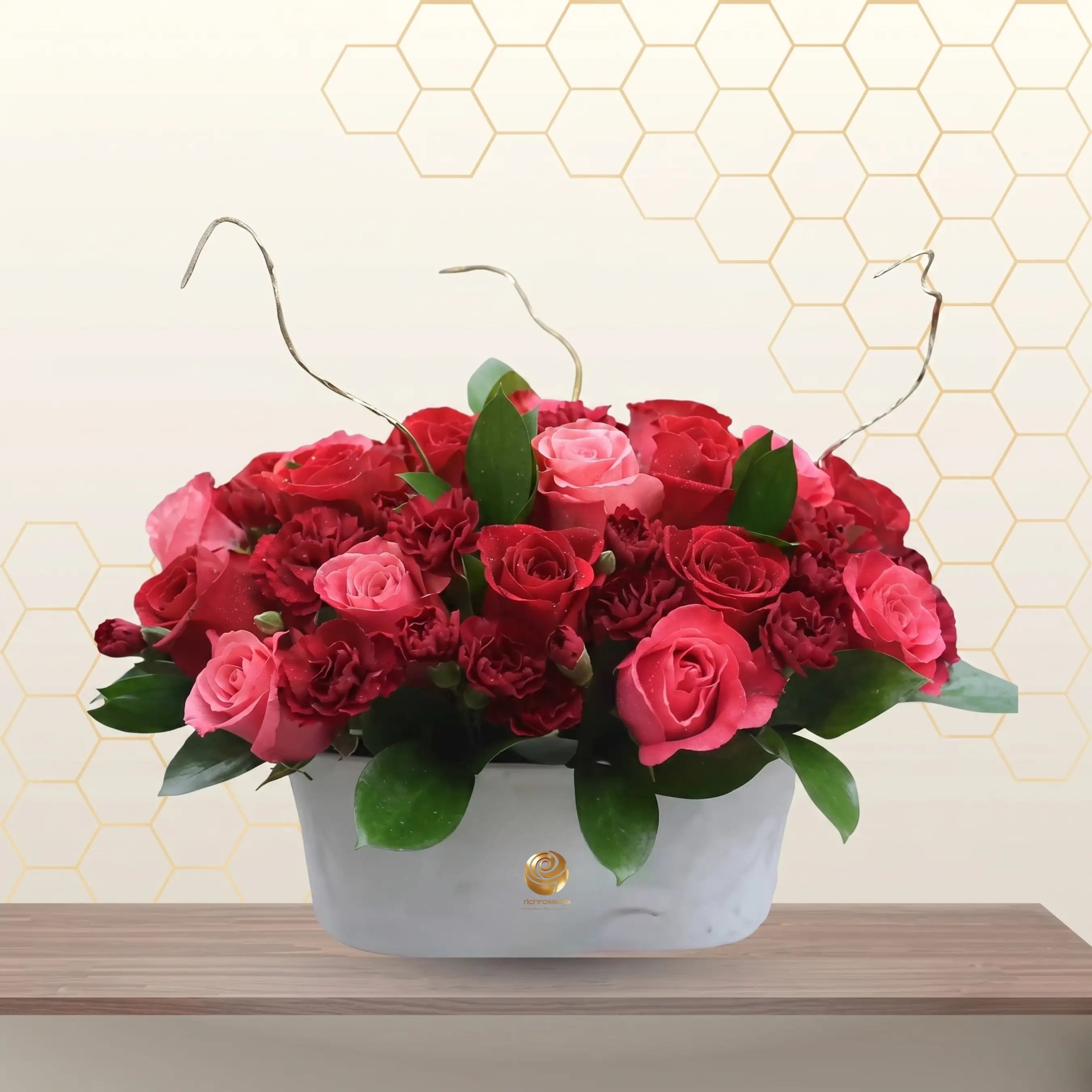 Nicola tabletop floral arrangement featuring red roses, pink roses, and red carnations in a white ceramic boat-shaped vase with decorative dry twigs. Main image