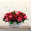 Nicola tabletop floral arrangement featuring red roses, pink roses, and red carnations in a white ceramic boat-shaped vase with decorative dry twigs.