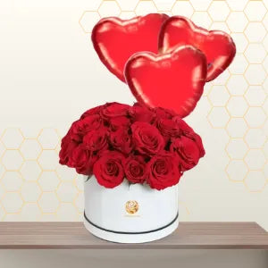 Round white hat box filled with red roses, with three red heart-shaped foil balloons floating behind it. Main image